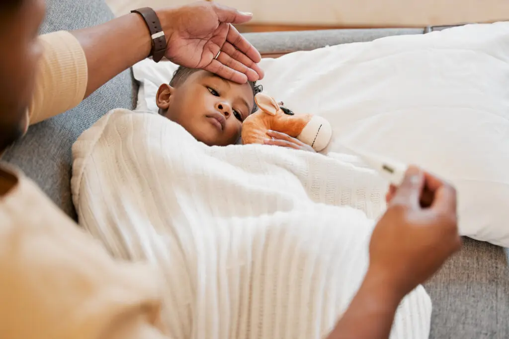 A child lying on a couch under a blanket while an adult checks their forehead for fever and holds a thermometer, with the child holding a stuffed toy.