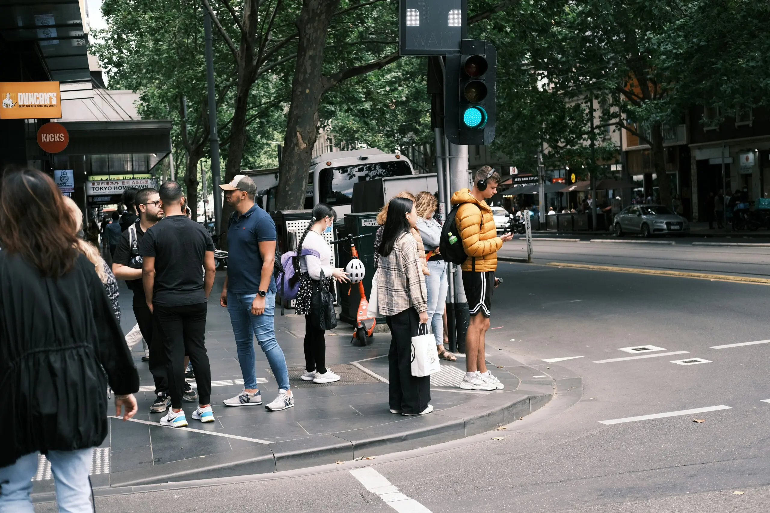 People standing on the sidewalk, engaged in conversation, with buildings and trees in the background.