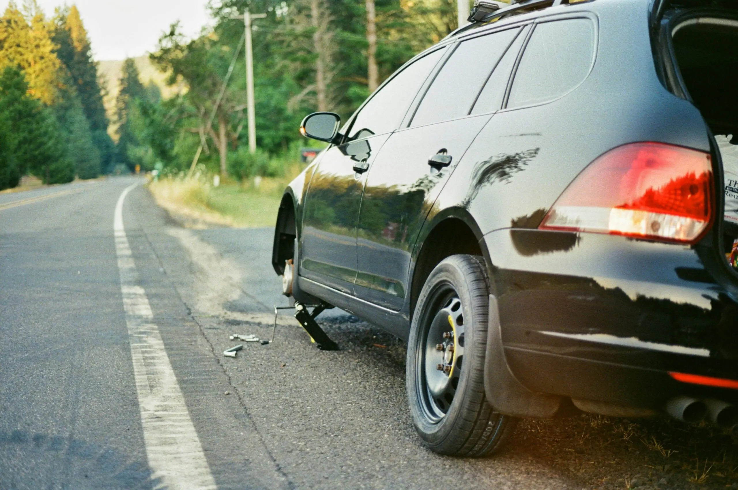 A black car parked on the side of a road, surrounded by greenery and a clear blue sky.