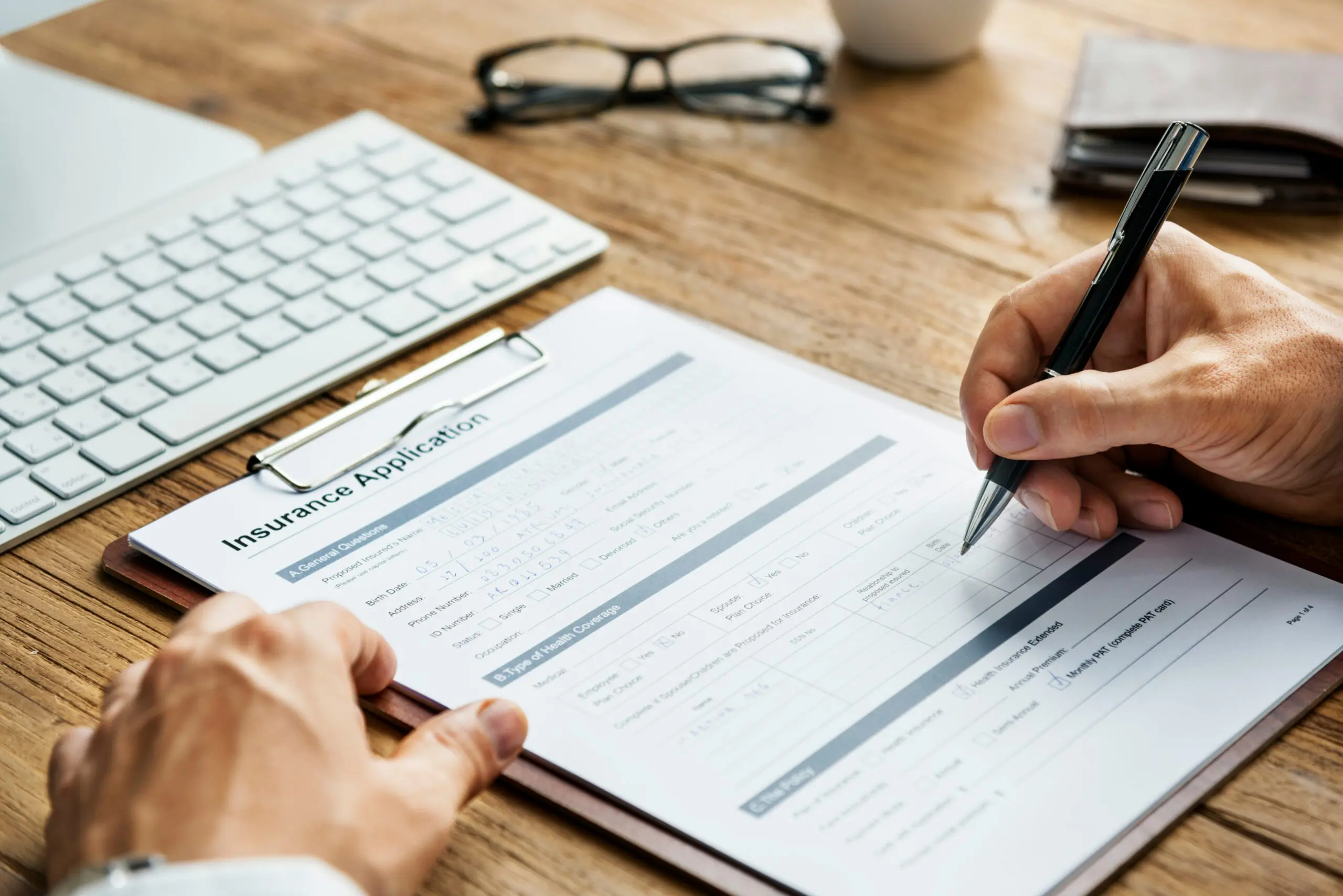Close-up of a person filling out an insurance application form on a clipboard at a desk, with a keyboard, glasses, and notebook nearby.