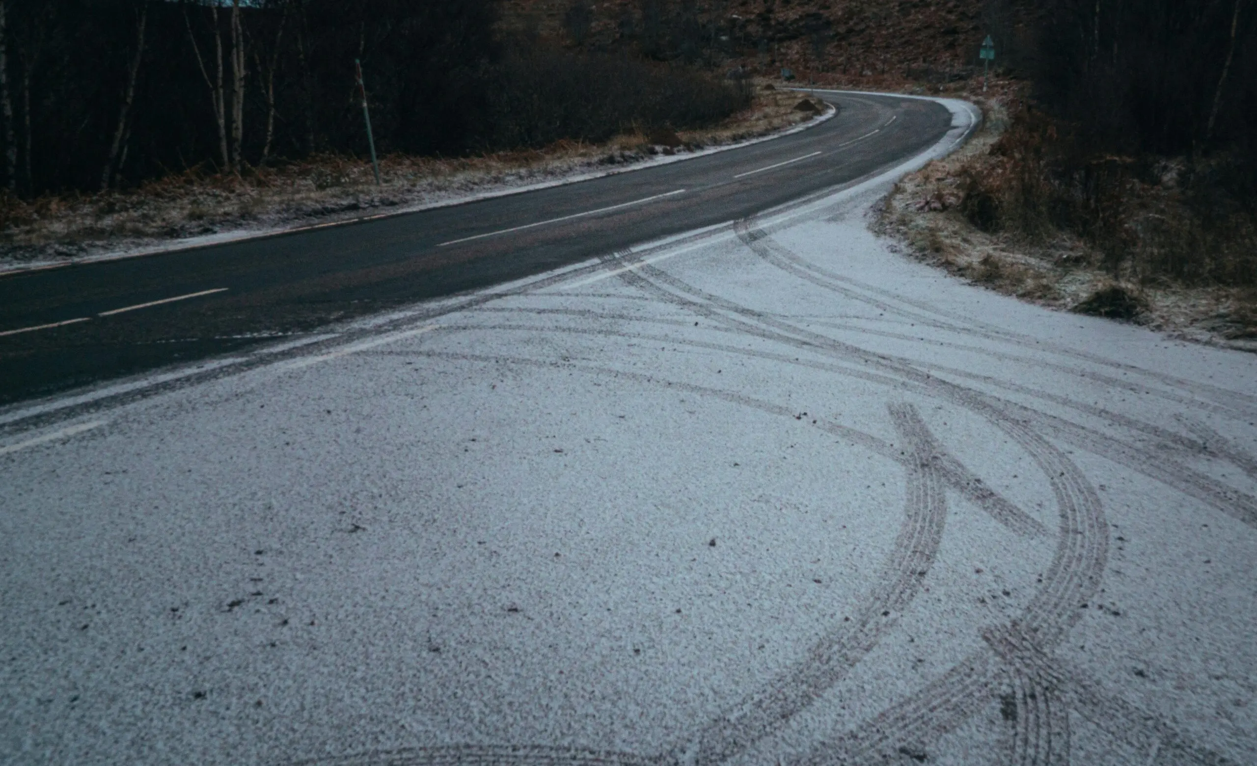 Curving road in a wooded area with a light layer of snow on the ground and visible tire tracks leading onto the roadway.