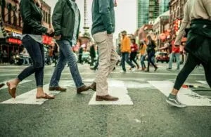 Pedestrians crossing a busy city street at a crosswalk, with people walking in both directions amid urban buildings and traffic in the background