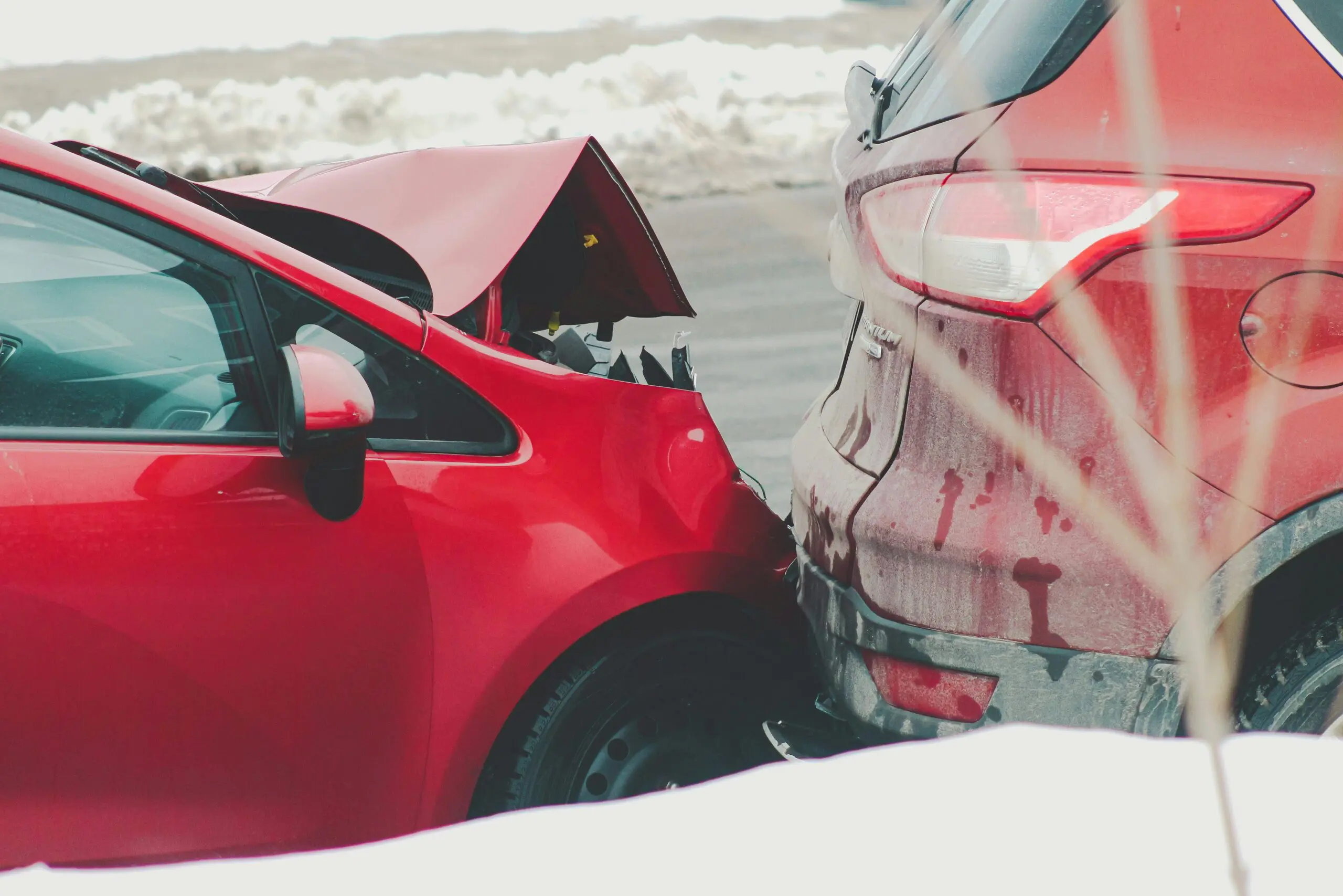 Rear-end car collision showing a red vehicle with a crumpled hood impacting the back of an SUV, with visible damage and debris on a cold, outdoor roadway