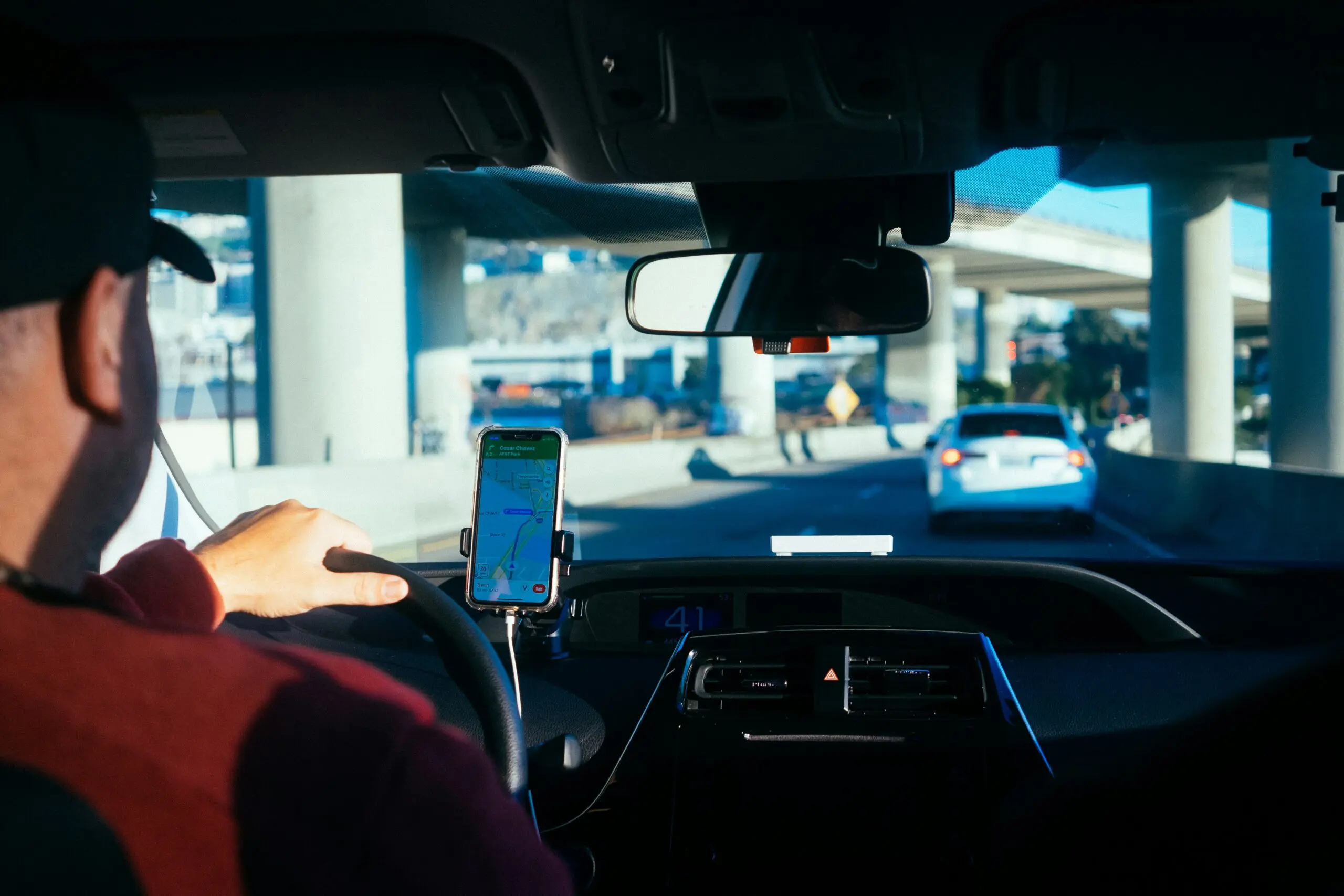 Driver using a smartphone navigation app mounted on the dashboard while driving on a highway, with traffic visible ahead and overpass structures in the background