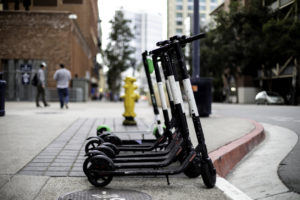 Several electric scooters lined up on a city sidewalk near a curb, with pedestrians and buildings blurred in the background.