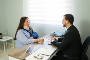 A man and woman engaged in conversation while seated at a desk, with papers and a laptop in front of them.
